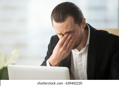 Frustrated businessman feels pain in eyes because of eyesight overstrain after long computer work. Tired young man massaging eyes in front of laptop. Eyes fatigue, headache or dizziness at workplace 