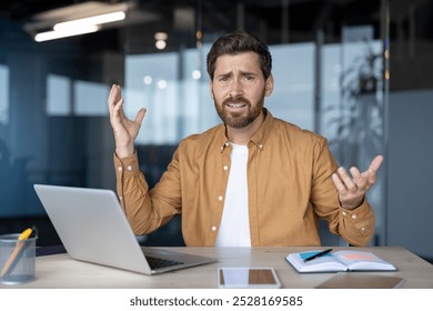 Frustrated businessman gestures at desk with laptop, notebook, phone, suggesting tech issue or confusion in office. Uncertainty and stress in workplace captured in moment. - Powered by Shutterstock