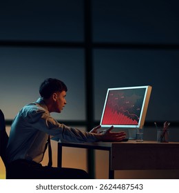 Frustrated businessman in office at night gestures angrily at computer screen showing dramatic financial collapse and red graph lines. Concept of business, crisis, decline, economy