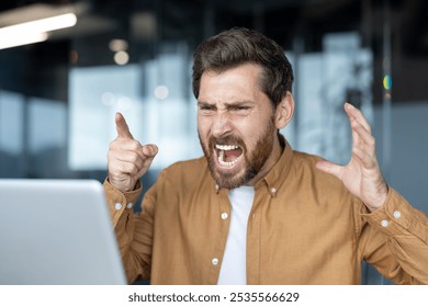 Frustrated businessman yelling at laptop during video call. Intense anger and stress evident in facial expressions. Office setting hints at workplace conflict, emotion, frustration, and tension. - Powered by Shutterstock