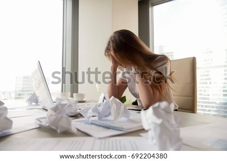 Frustrated businesswoman holding head in hands sitting at office desk covered with crumpled paper, feeling tired after unproductive work, giving up, creative crisis, no motivation, lack of ideas