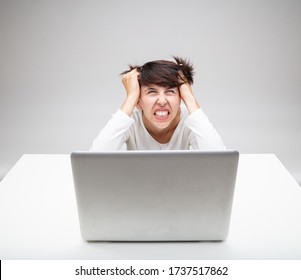 Frustrated businesswoman tearing at her hair in exasperation as she sits working at a laptop on a white desk against a grey background