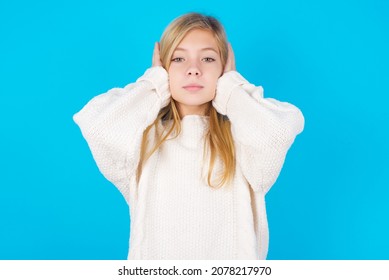 Frustrated Caucasian Little Kid Girl Wearing White Knitted Sweater Over Blue Background Plugging Ears With Hands Does Not Wanting To Listen Hard Rock, Noise Or Loud Music.