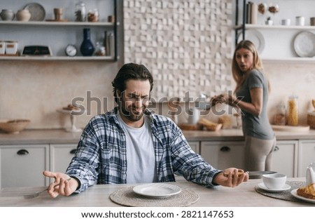 Frustrated Caucasian Man At Kitchen Table With Woman Behind During Tense Situation, Conflict