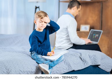 Frustrated Child. Pleasant Little Boy Holding A Drawing Book And Touching His Forehead, Struggling To Come Up With New Ideas About New Picture While His Father Working