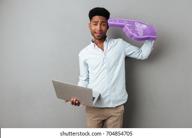 Frustrated Confused African Man Holding Laptop Computer And Wearing Foam Finger Isolated Over Gray Background