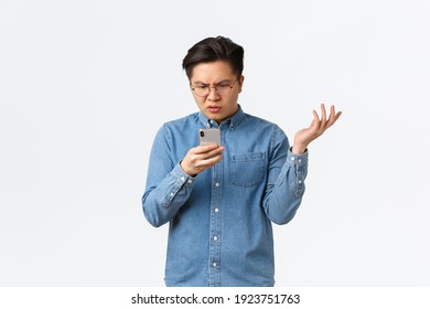 Frustrated And Confused Asian Guy In Glasses Looking Puzzled At Mobile Phone Screen, Raising Hand Up Questioned, Cant Understand What Happening Online, Standing White Background Perplexed