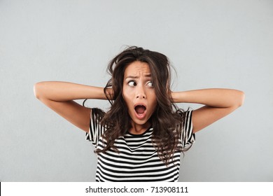 Frustrated confused asian woman keeping hands in her hair and screaming isolated over gray background