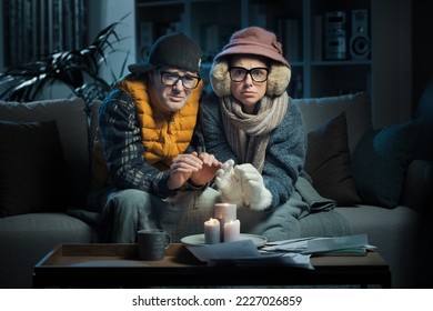 Frustrated couple sitting on the couch and feeling cold at home, they are trying to save money on their utility bills