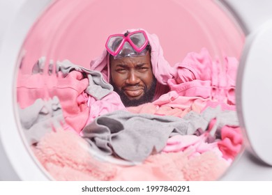 Frustrated dark skinned adult man cries from despair wears snorkeling mask covered with pile of laundry sticks head in washing machine drum. Automatic self service laundromat. Laundering at home