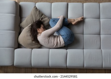 Frustrated depressed millennial girl lying in embryo pose on couch, sleeping, drowsing, feeling stress, apathy, suffering from abdominal pain, bellyache. Depression concept. Top view