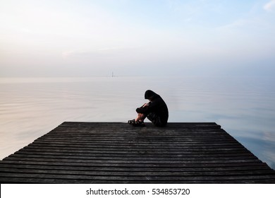 Frustrated depression man in the hood sitting on wooden bridge, near the beach on sunset. Concept of Major depressive disorder, unemployed, sadness, depressed and human problems
