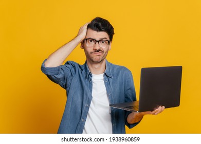 Frustrated displeased unshaven caucasian guy in glasses and in denim shirt, holds a laptop, looks confusedly at the camera, holding his head, without mood, stands on an isolated orange background - Shutterstock ID 1938960859