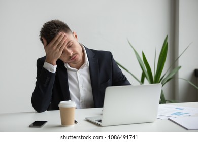 Frustrated fatigued millennial businessman having strong headache tired from laptop use touching forehead, stressed overworked company ceo suffers from migraine at work, aching head in office concept