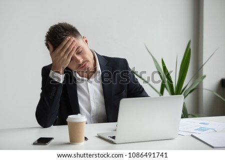 Frustrated fatigued millennial businessman having strong headache tired from laptop use touching forehead, stressed overworked company ceo suffers from migraine at work, aching head in office concept