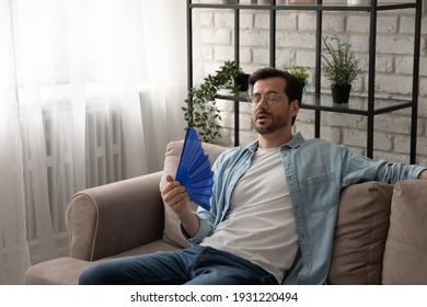 Frustrated Guy Recline On Sofa Suffer From Too Hot Temperature At Home Breath Hard With Dehydrated Air Cool Down With Hand Fan. Tired Young Male Complain On Extra Heat At Living Room Feel Bad Sweaty