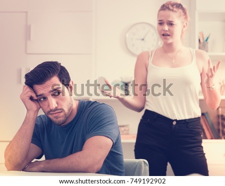 Frustrated guy sitting at home table on background with dissatisfied girlfriend