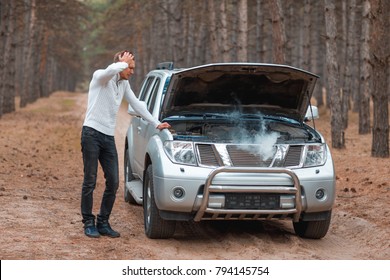 A Frustrated Guy, Standing Near A Broken Car With An Open Hood With Smoke, Stands And Holds Head.