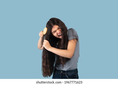 Frustrated Indian Teen Girl Trying To Brush Her Tangled Long Locks On Blue Studio Background. Pretty Adolescent Having Bad Hair Day, Upset Over Messy Hairdo. Hairdressing And Beauty Concept