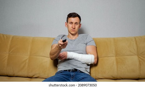 Frustrated Man With Broken Spoke-bone In Gypsum Band Switches TV On Using Remote Controller Sitting On Comfortable Sofa In Living Room.