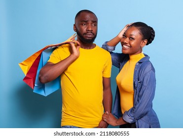 Frustrated man carrying shopping bags for cheerful girlfriend. Shopaholic woman making purchase from retail store center, buying clothes while her boyfriend feels frustrated and tired. - Shutterstock ID 2118080555