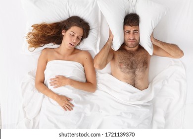 Frustrated Man Covers Ears With Pillow, Cannot Fall Asleep Because Of Loud Wife Snoring, Suffers From Insomnia, Pose In Bedroom. Tired Man Irritated By Womans Snoring. People, Health, Sleep Disorder