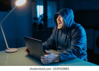 A frustrated man in a hoodie sits with his laptop in a dark room, displaying his anger and stress after experiencing failure in his hacking efforts. - Powered by Shutterstock