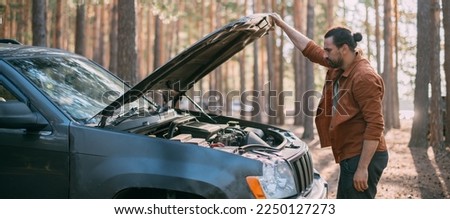 A frustrated man near a broken car with an open hood far outside the city in the woods. The car broke down while traveling. A young guy looks under the hood of a car.