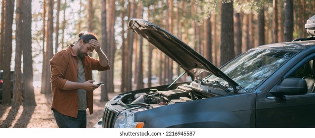 A Frustrated Man With A Phone In His Hands Near A Broken Car With An Open Hood Far Outside The City In The Woods. The Car Broke Down While Traveling. A Young Guy Looks Under The Hood Of A Car.