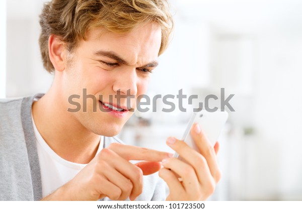 Frustrated man sending text message using mobile\
phone at home