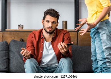 Frustrated Man Sitting On Sofa And Showing Shrug Gesture Near Angry Girlfriend