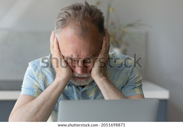 Frustrated middle aged older retied man in\
eyeglasses covering ears, feeling stressed using computer software\
applications, considering financial problem, suffering from lack of\
money managing\
budget.