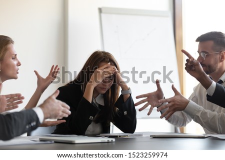 Frustrated millennial female worker sitting at table with colleagues, felling tired of working quarreling at business meeting. Upset stressed young businesswoman suffering from head ache at office.