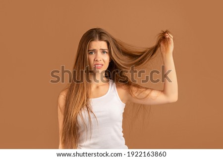 Frustrated millennial woman showing her damaged long locks on brown studio background. Pretty young lady having bad hair day, upset over her messy hairdo. Hairdressing services concept
