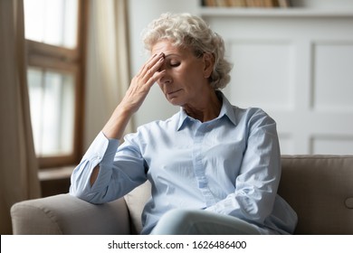 Frustrated Nervous Middle Aged Hoary Woman Sitting On Sofa, Touching Forehead, Suffering From Headache Alone At Home. Worried Stressed Mature Older Grandmother Having Painful Feelings In Head.