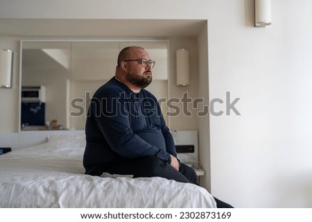 Frustrated overweight man sits on bed sadly looking to window feeling depression from difficult relations, works trouble, health problem. Unhappy male think about hopelessness need psychological help 