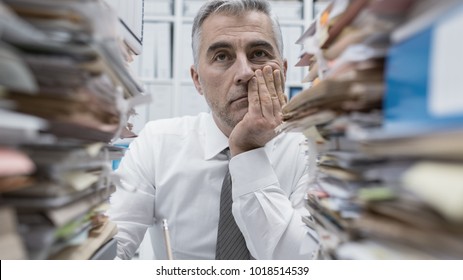 Frustrated overwhelmed executive working in the office and overloaded with paperwork, he is leaning on his arm and feeling depressed