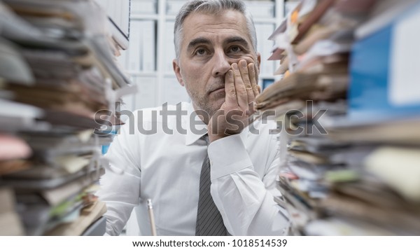 Frustrated overwhelmed executive working in the\
office and overloaded with paperwork, he is leaning on his arm and\
feeling depressed