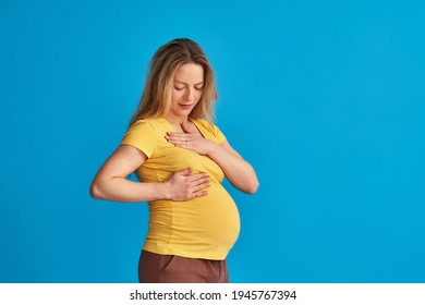 A frustrated pregnant woman checks her breasts for a seal while standing on a blue studio background. Chest pain, neoplasm and discharge from the chest. Mastopathy of pregnant women. Copy space.
