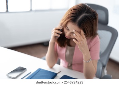 A frustrated professional woman sits at her office desk, holding her head while on a phone call. An image conveys workplace stress, problem-solving, and handling challenges in a corporate environment - Powered by Shutterstock