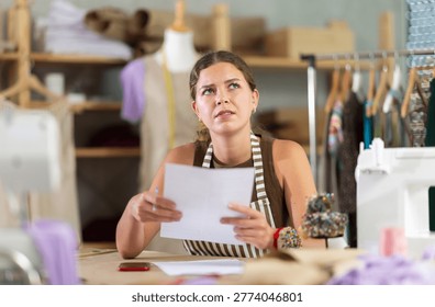 Frustrated and puzzled dressmaker with documents sits at a table in a sewing workshop. Seamstress makes notes on paper and calculates the budget for sewing clothes