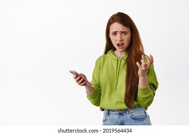 Frustrated redhead girl complains at something on mobile phone, look annoyed and holding smartphone, frowning disappointed, white background