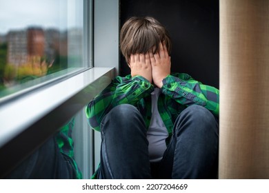 A frustrated, sad and lonely teenager of 12 years old is sitting on the windowsill of the house, covering his face with his hands. Children's social psychology and emotions. - Powered by Shutterstock