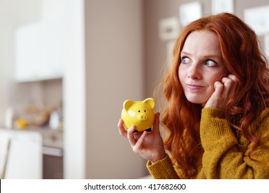 Frustrierte Einzelfrau in rotem Haar und gelbem Pullover, die ein wenig gelbes Sparschwein hält, während sie wegschaut