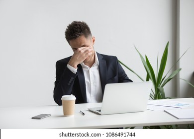 Frustrated stressed businessman feeling tired of computer work sitting at workplace, exhausted man in suit suffers from eye strain or blurry vision problem after long laptop use, eyes fatigue concept