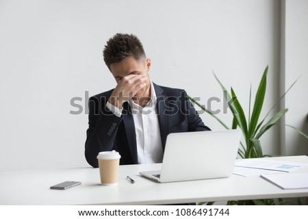 Frustrated stressed businessman feeling tired of computer work sitting at workplace, exhausted man in suit suffers from eye strain or blurry vision problem after long laptop use, eyes fatigue concept