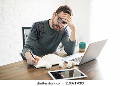 Frustrated And Stressed Male Writer Sitting With Laptop And Writing In A Book