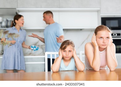 Frustrated teenage girls covering ears while sitting at table in home kitchen, unwilling to listen to mother and father quarrel. Parental conflict concept - Powered by Shutterstock