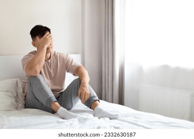 Frustrated unhappy depressed middle aged asian man wearing pajamas sitting alone on bed at home, covering his face, experiencing midlife crisis, going through divorce, copy space - Powered by Shutterstock