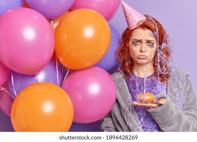 Frustrated unhappy redhead Caucasian woman with spoiled makeup sad to be lonely on her birthday holds cupcake with burning candle bunch of inflated balloons has party at home wears cone hat on head